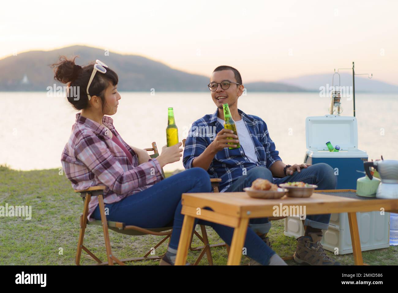 Ein asiatisches Paar trinkt Bier aus einer Flasche in seinem Campingbereich mit See im Hintergrund während des Sonnenuntergangs. Stockfoto