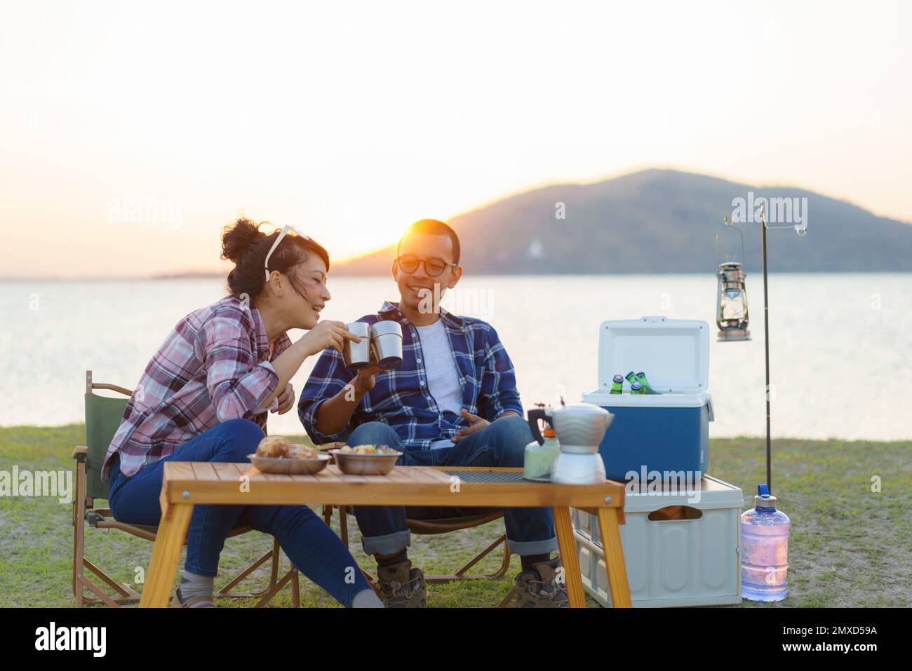 Ein asiatisches Paar trinkt eine Tasse Kaffee oder Wasser auf dem Campingplatz mit See im Hintergrund bei Sonnenuntergang Stockfoto