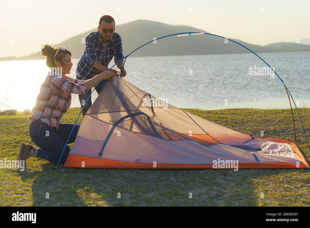 Ein asiatisches Paar bereitet ein Zelt vor, um auf dem Rasen zu campen, während der See im Hintergrund bei Sonnenuntergang liegt Stockfoto