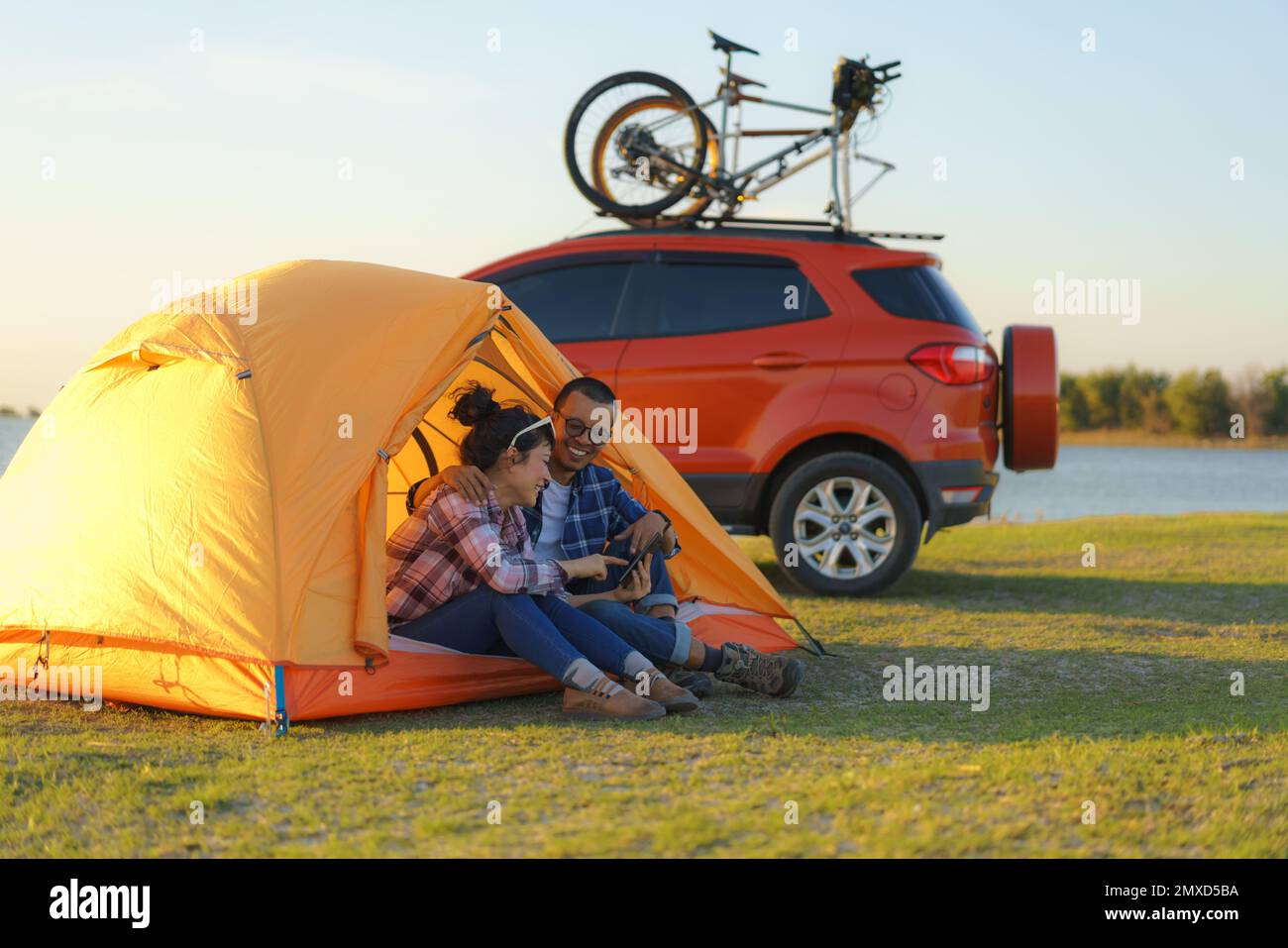 Ein junges asiatisches Paar, das Selfie mit einer Smartphone-Kamera macht, indem es in einem Zelt sitzt und mit dem See im Hintergrund bei Sonnenuntergang zeltet Stockfoto