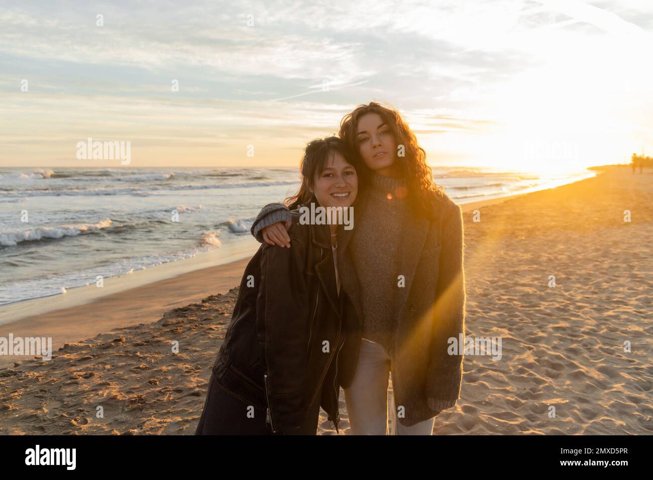 Lockige Frau, die eine lächelnde Freundin am Strand während des Sonnenuntergangs in Barcelona umarmt Stockfoto