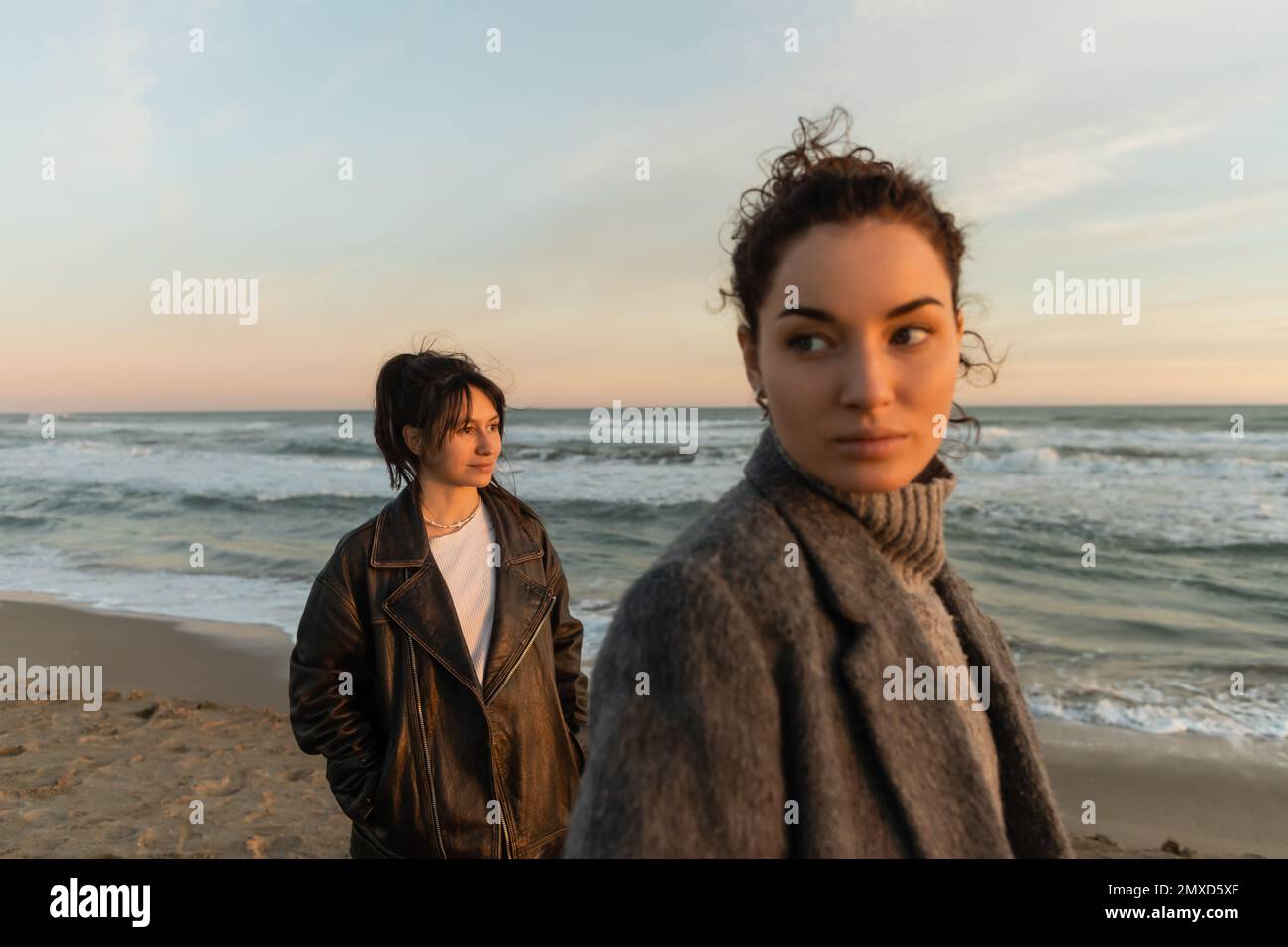 Lächelnde Frau, die in der Nähe eines verschwommenen Freundes am Strand während des Sonnenuntergangs in Barcelona wegschaut Stockfoto