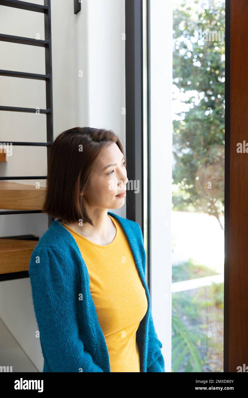 Vertikal einer durchdachten asiatischen Frau, die steht, aus dem Wohnzimmerfenster schaut, Kopierraum Stockfoto