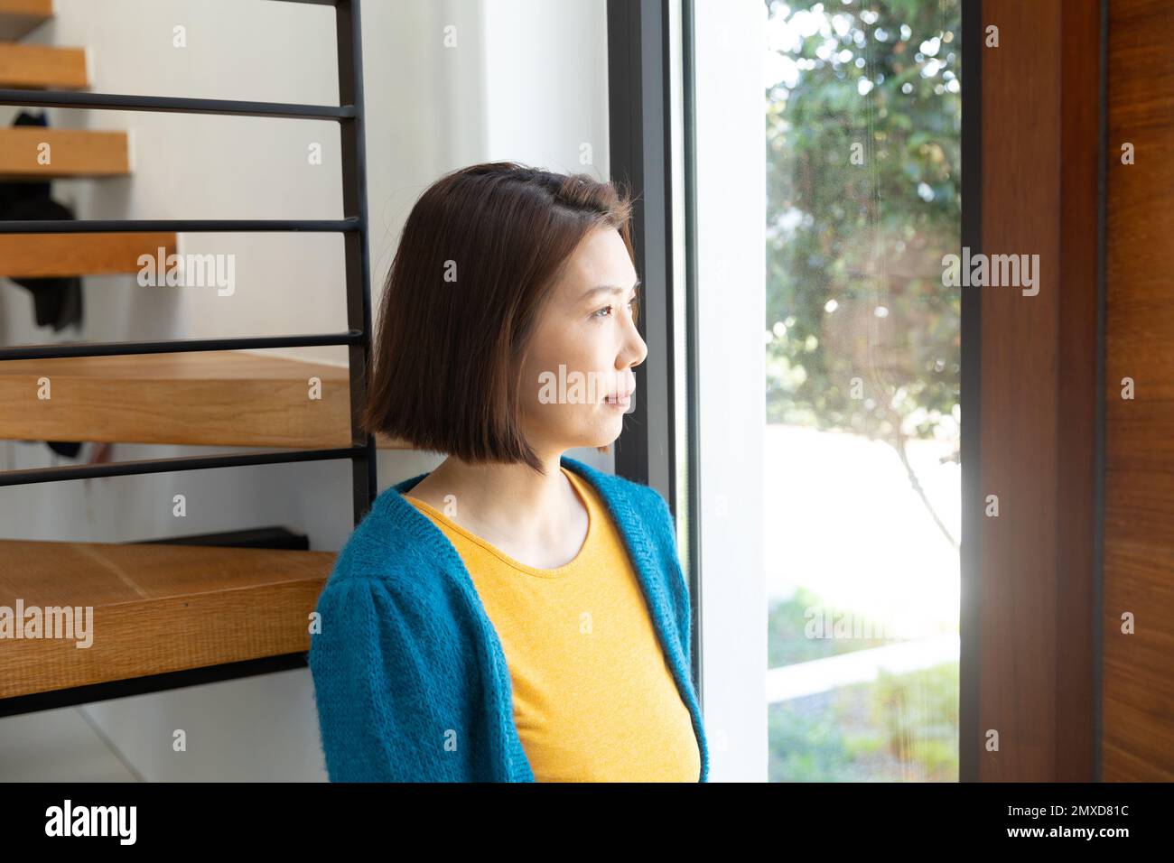 Rücksichtsvolle asiatische Frau steht, schaut aus dem Wohnzimmerfenster, Kopierraum Stockfoto