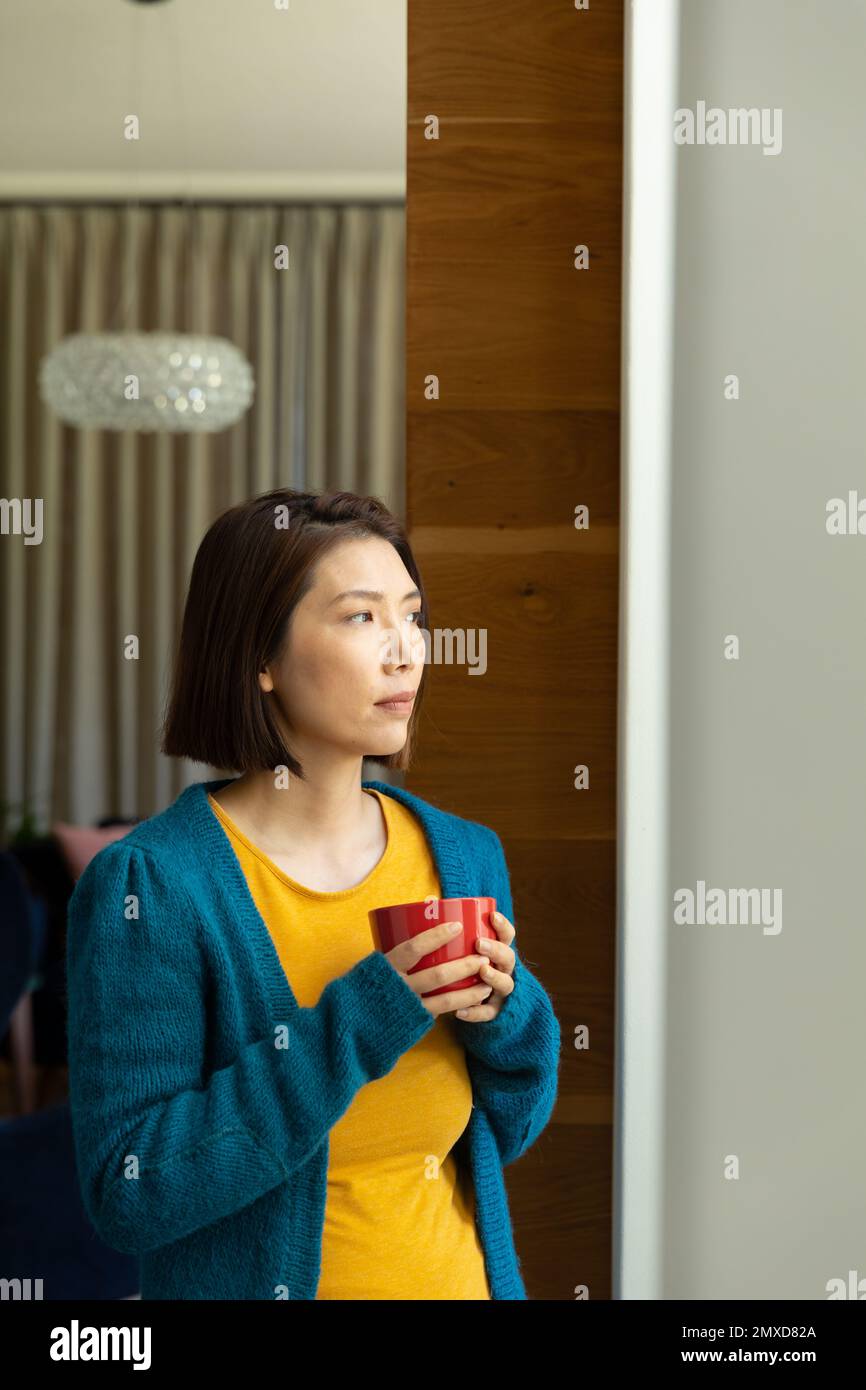 Eine durchdachte asiatische Frau mit einer Tasse Kaffee, die aus dem Wohnzimmerfenster blickt, einen Kopierraum Stockfoto