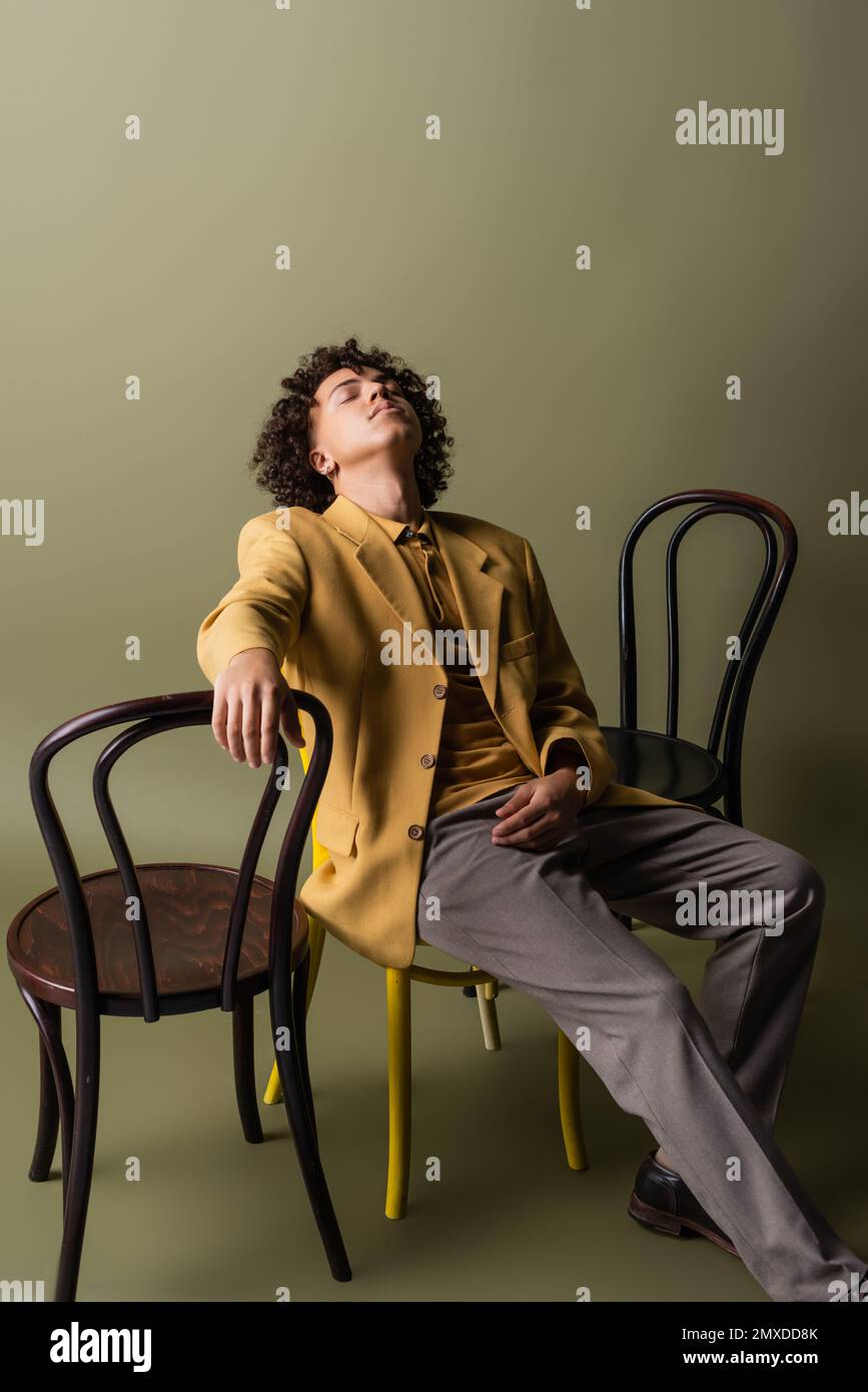 Junger und trendiger afroamerikanischer Mann mit geschlossenen Augen, der neben schwarzen Stühlen auf olivgrauem Hintergrund sitzt, Stockbild Stockfoto