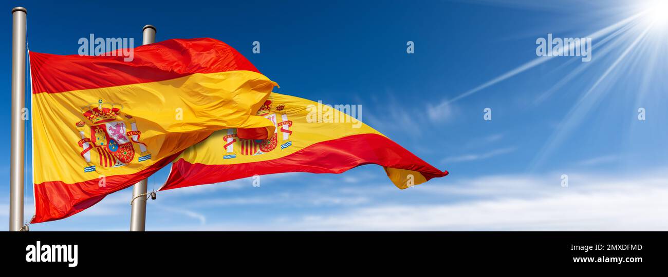 Nahaufnahme von zwei spanischen Nationalflaggen (La Rojigualda) mit Fahnenstange, die im Wind an einem blauen Himmel mit Wolken, Sonnenstrahlen und Kopierraum weht. Stockfoto