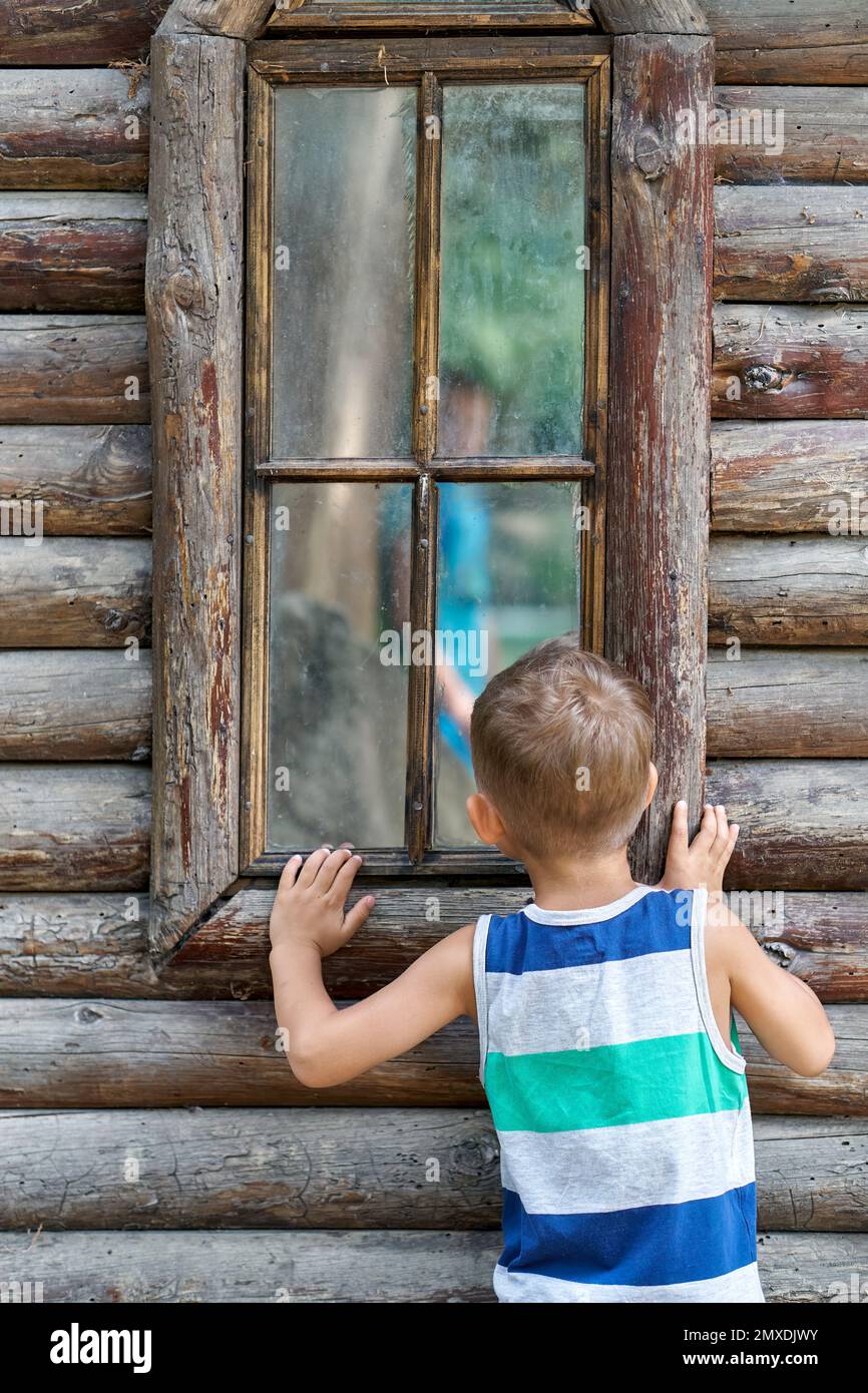Aufgeregter Vorschuljunge schaut in das Fenster des alten Holzhauses. Ein neugieriges Kind sieht sich Reflexionen auf Fensterglas an und möchte das Reiseziel erkunden Stockfoto