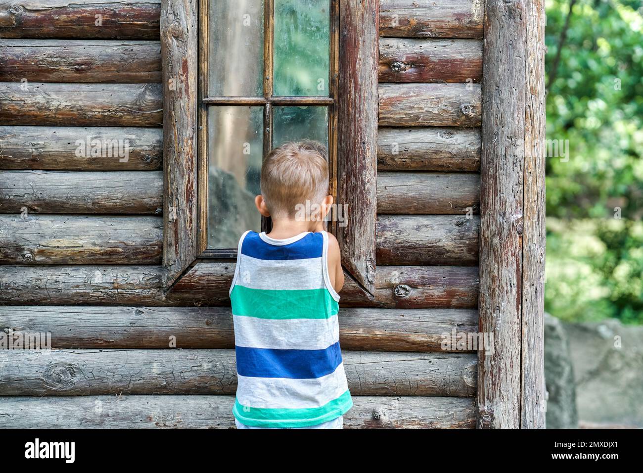 Aufgeregter Vorschuljunge schaut in das Fenster des alten Holzhauses. Ein neugieriges Kind sieht sich Reflexionen auf Fensterglas an und möchte das Reiseziel erkunden Stockfoto