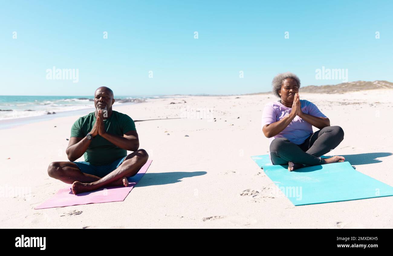 afroamerikanisches Seniorenpaar mit gekreuzten Beinen am Strand vor klarem Himmel zu meditieren Stockfoto