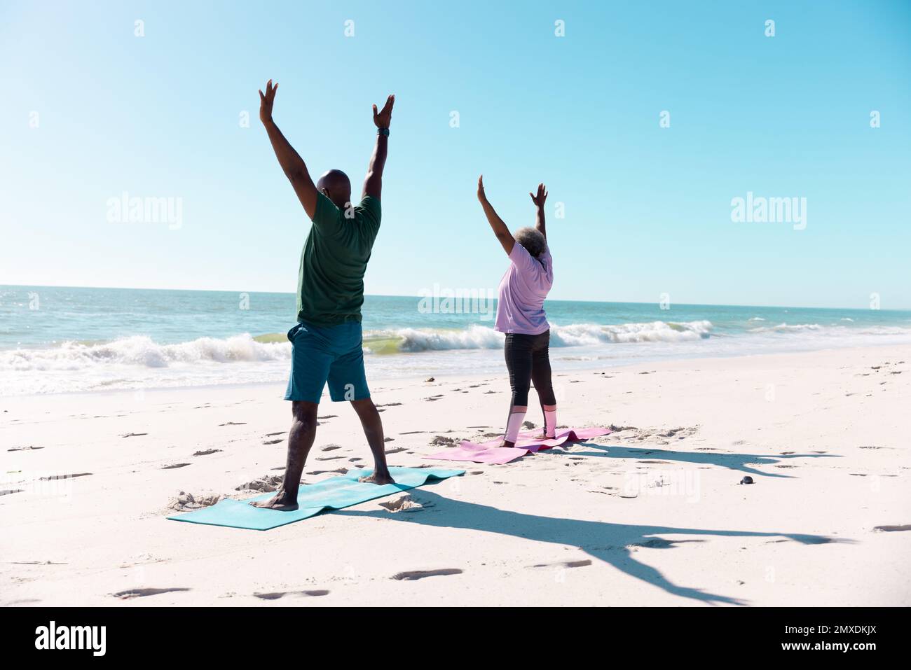 afroamerikanisches Seniorenpaar mit erhobenen Armen beim Training auf Matten am Strand unter einer klaren blauen Sackgasse Stockfoto