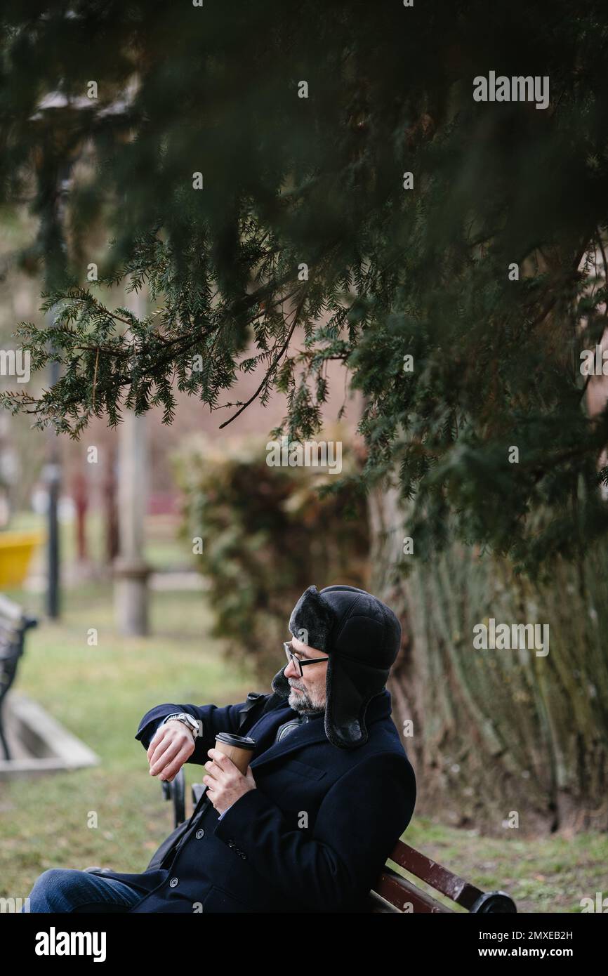 Ein älterer Mann genießt einen kalten Wintermorgen in der Stadt. Ein Mann sitzt auf einer Bank auf dem Platz, das Konzept von Frieden und Vitalität. Stockfoto
