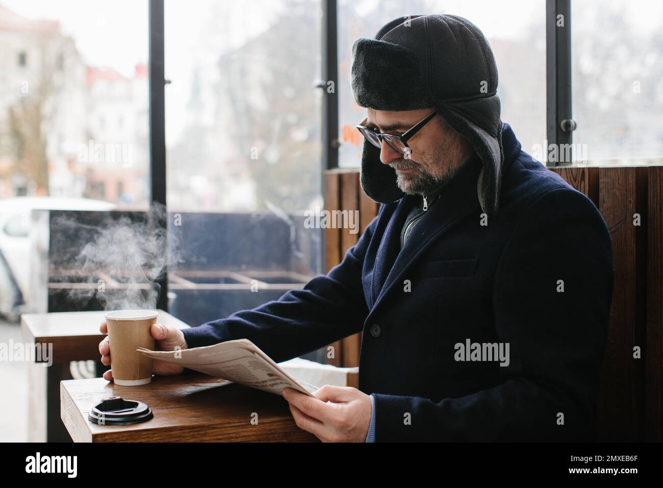 Ein grauhaariger Mann in warmen Kleidern liest eine frische Zeitung und trinkt heißen Tee oder Kaffee auf der Straße. Stockfoto