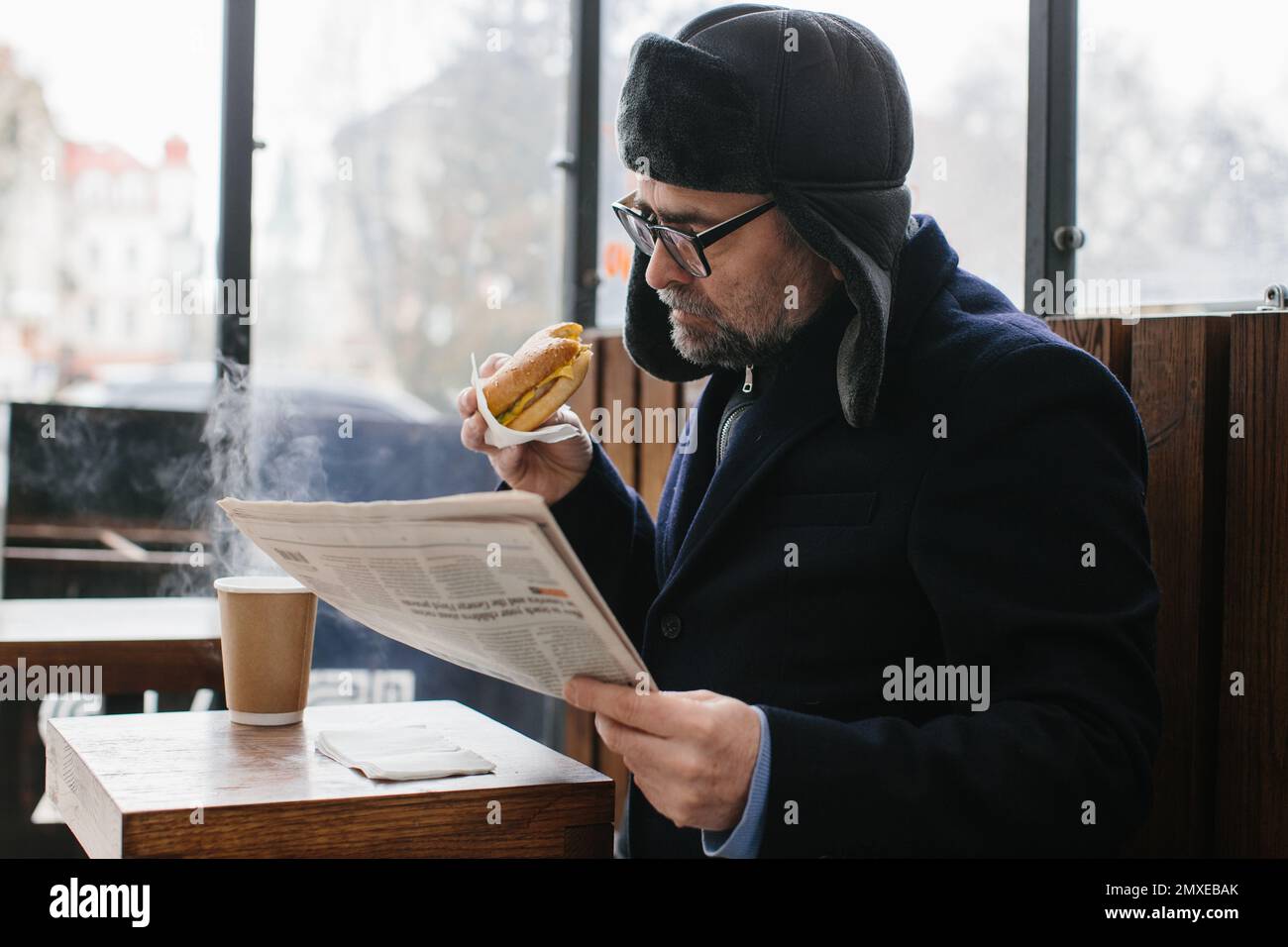 Ein bärtiger Mann mittleren Alters isst einen leckeren Burger und liest im Winter auf einer Stadtstraße die Zeitung von heute. Street Food-Konzept. Stockfoto