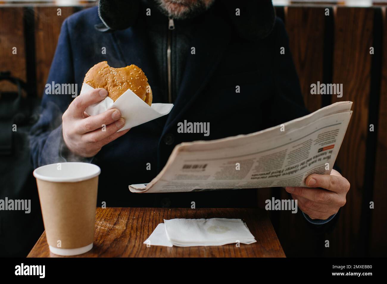 Ein bärtiger Mann mittleren Alters isst einen leckeren Burger und liest im Winter auf einer Stadtstraße die Zeitung von heute. Street Food-Konzept. Stockfoto