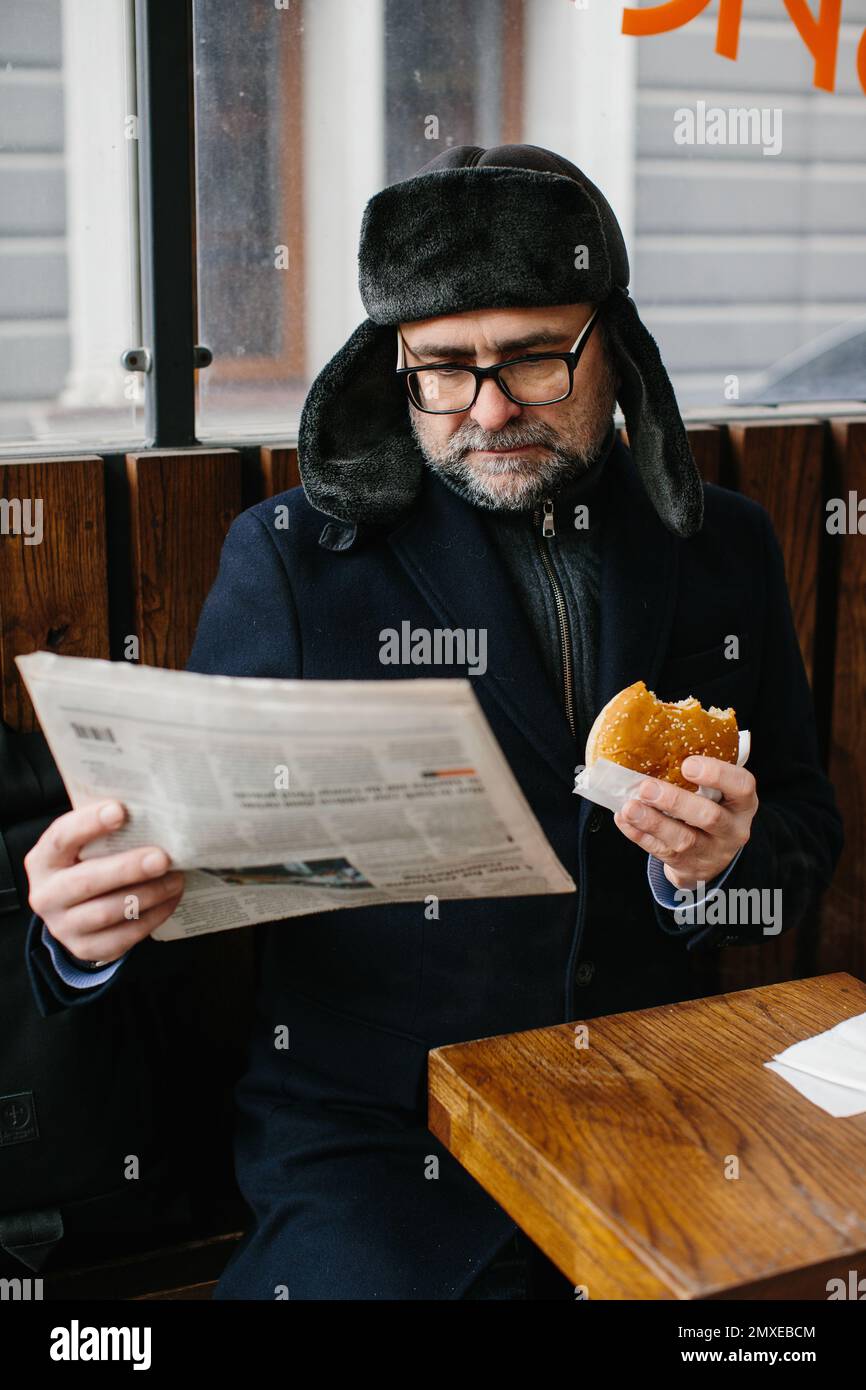 Ein bärtiger Mann mittleren Alters isst einen leckeren Burger und liest im Winter auf einer Stadtstraße die Zeitung von heute. Street Food-Konzept. Stockfoto