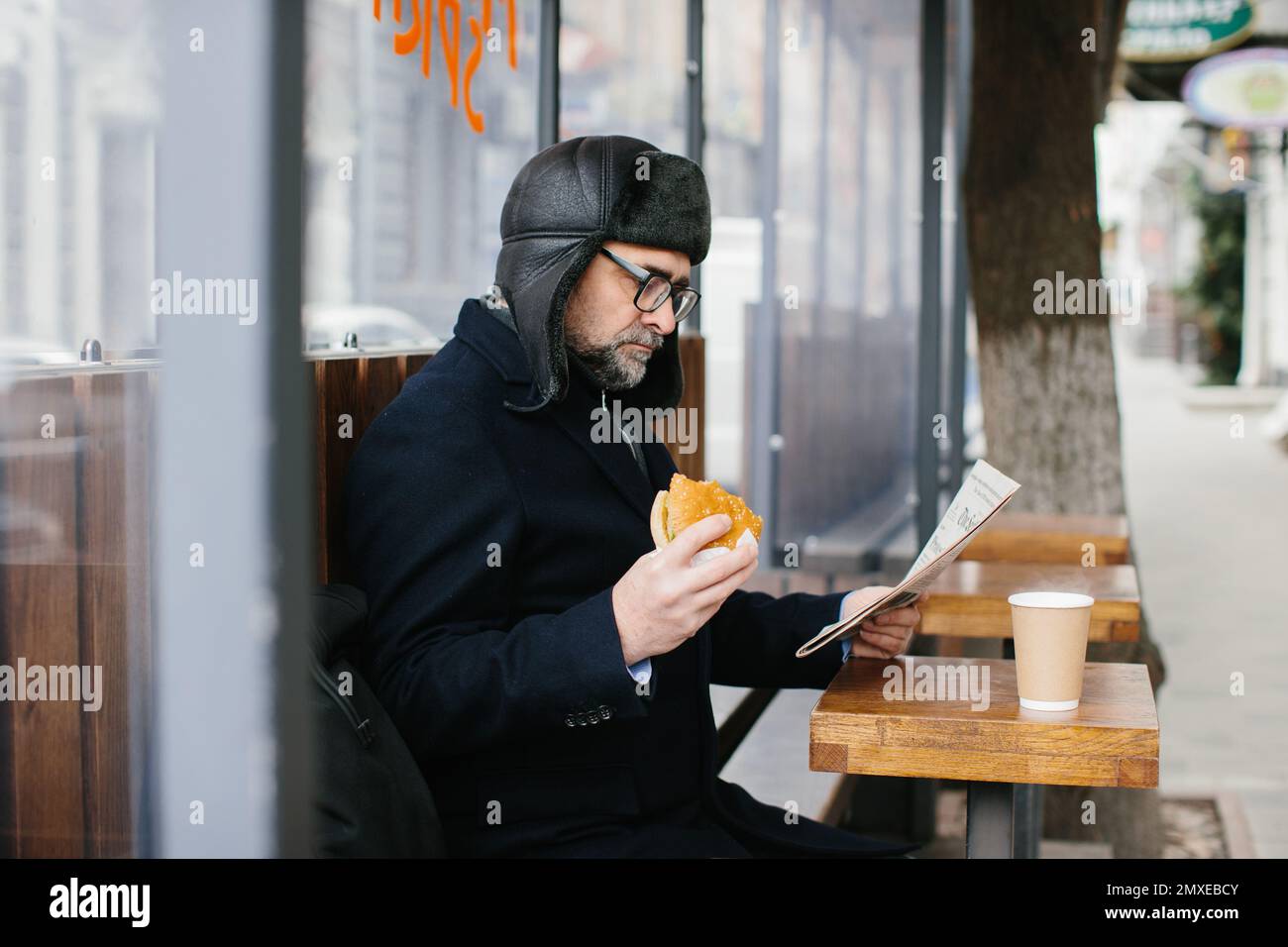 Ein bärtiger Mann mittleren Alters isst einen leckeren Burger und liest im Winter auf einer Stadtstraße die Zeitung von heute. Street Food-Konzept. Stockfoto