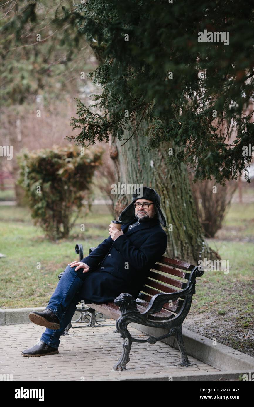 Ein älterer Mann genießt einen kalten Wintermorgen in der Stadt. Ein Mann sitzt auf einer Bank auf dem Platz, das Konzept von Frieden und Vitalität. Stockfoto