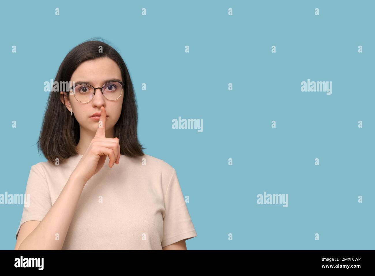 Porträt einer seriösen Schülerin mit Brille, die ihren Mund mit dem Finger auf blauem Hintergrund bedeckt. Das Mädchen will nicht reden. Nonverbale Kal Stockfoto