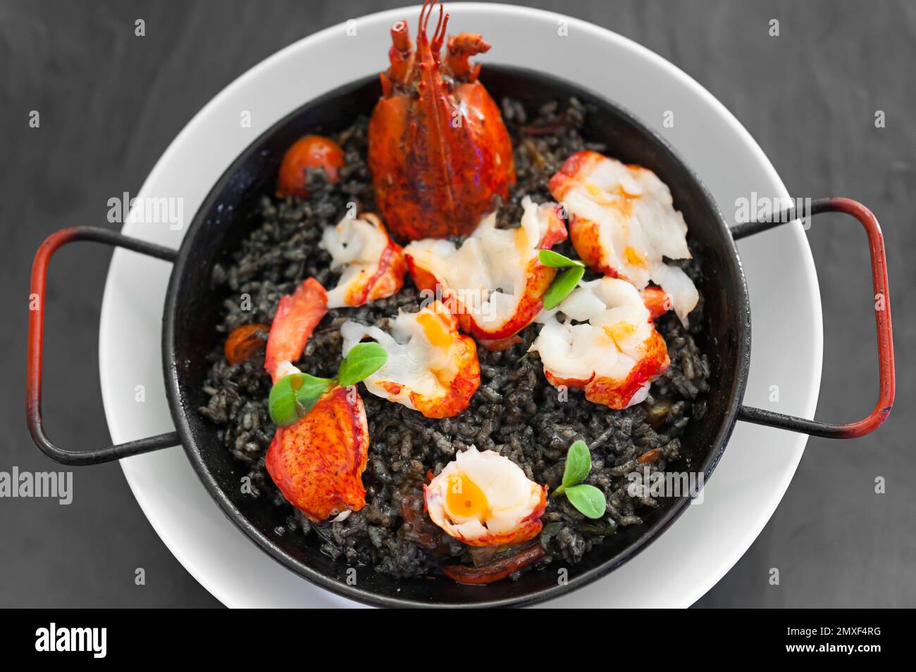 Solo-Paella mit Hummerscheiben, Tomatenstückchen, Tintenfischsauce, Kräutern, Safran. Heißes Gericht auf weißem Teller. Essen auf einem schwarzen Tisch. Moderne Verpflegung. Stockfoto