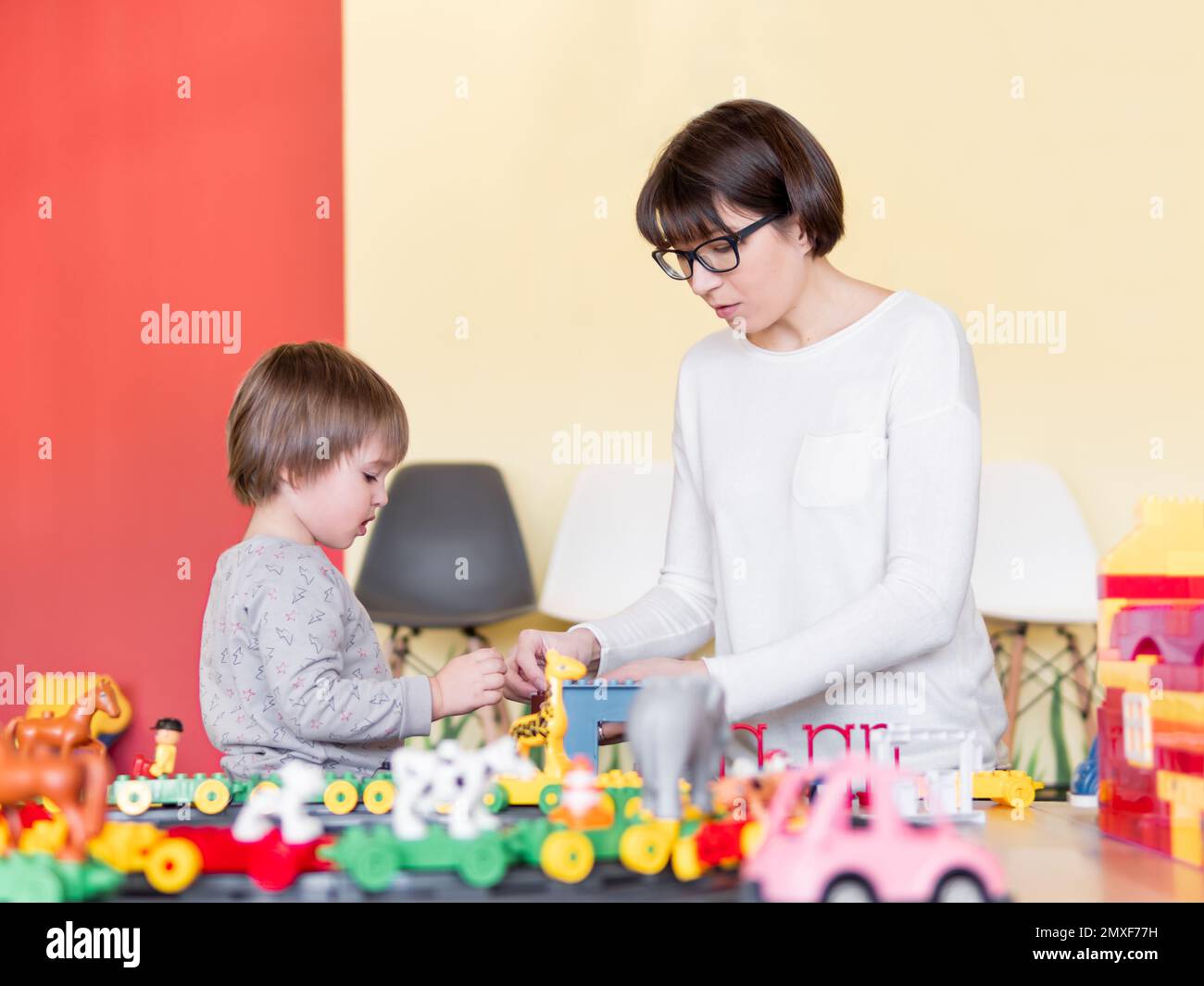 Ihr Kind spielt mit bunten Spielwürfeln, während seine Mutter oder sein Babysitter spielt. Der kleine Junge starrt auf den Spielzeugbauer. Das Innere des Kindergartens. Stockfoto
