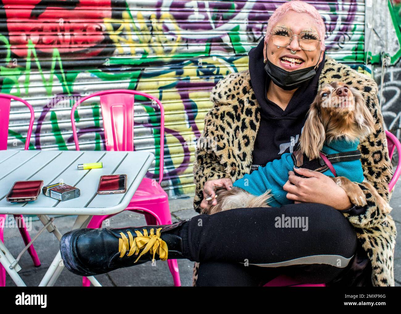 Weibliche Straßenhändlerin mit rosafarbenen Haaren trägt einen Mantel mit Leopardenmuster, Doc Marten Stiefel hält einen Hund, lächelt in die Kamera. Camden Markt Stockfoto