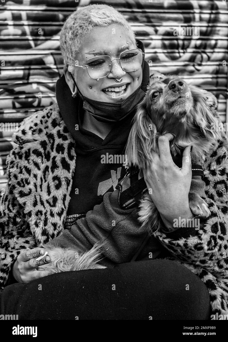 Weibliche Straßenhändlerin mit pinkfarbenen Haaren trägt einen Mantel mit Leopardenmuster, Doc Marten Stiefel hält einen Hund und lächelt in die Kamera. Camden Markt Stockfoto