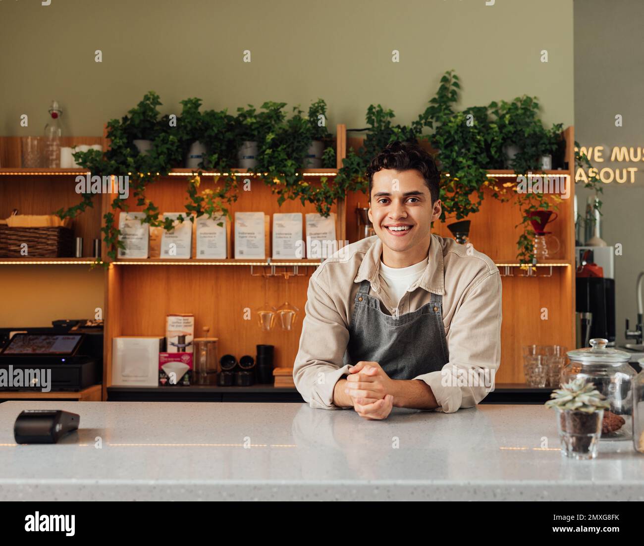 Lächelnder Coffee Shop-Besitzer in der Schürze lehnt sich an die Theke. Der Barkeeper aus dem Nahen Osten schaut in die Kamera. Stockfoto