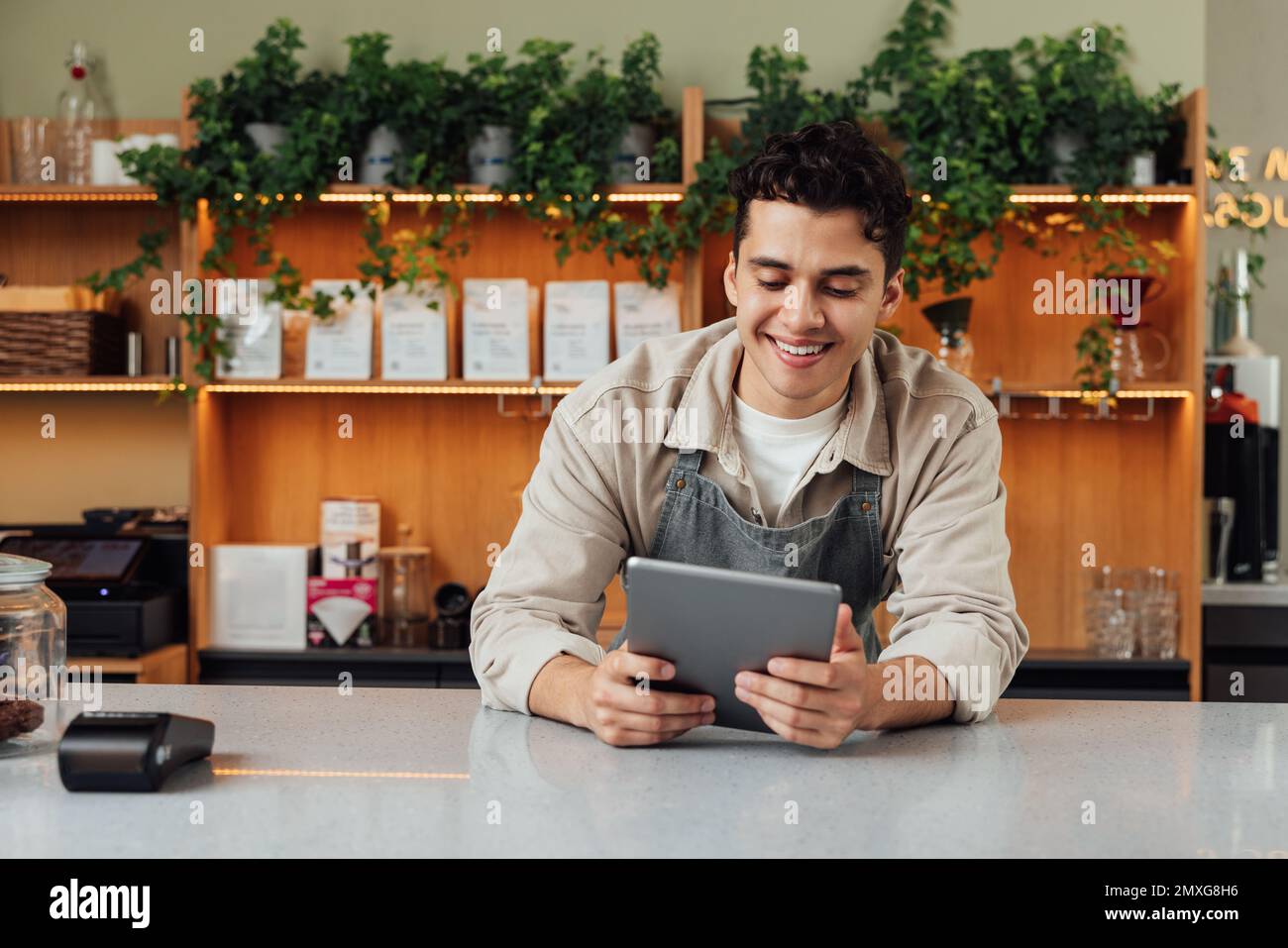 Lächelnder Besitzer eines Cafés am Tresen mit digitalem Tablet. Männlicher Barkeeper in einer Schürze, der auf einen tragbaren Computer schaut. Stockfoto