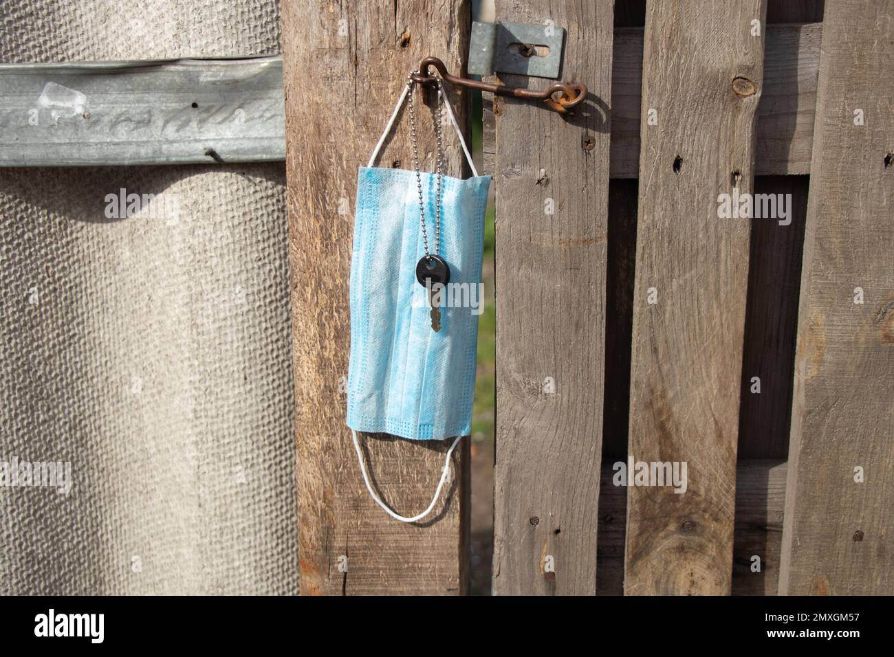 Medizinische Maske hängt an einem Haken und einem Türschlüssel an einem alten Holztor in der Ukraine auf der Straße währenddessen Stockfoto