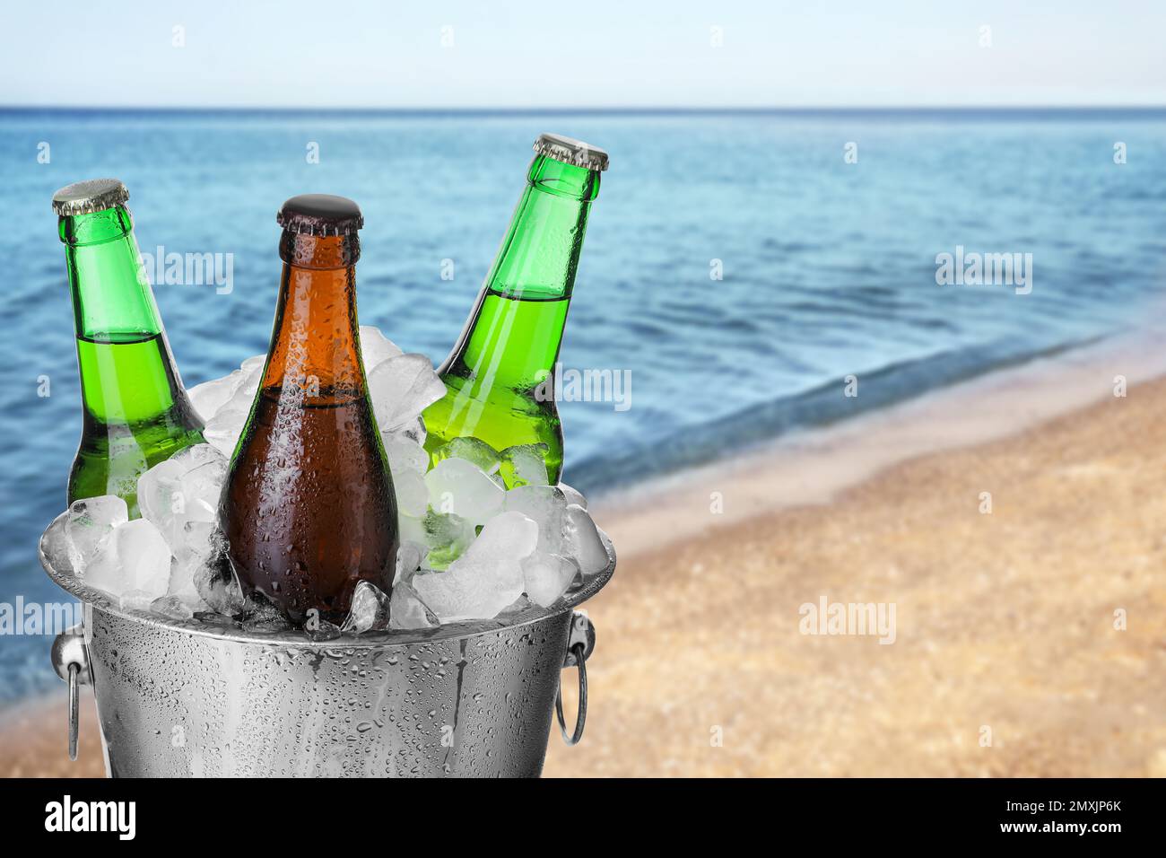 Flaschen Bier mit Eiswürfeln in einem Metalleimer gegen verschwommenes Meer und Sandstrand. Platz für Text Stockfoto