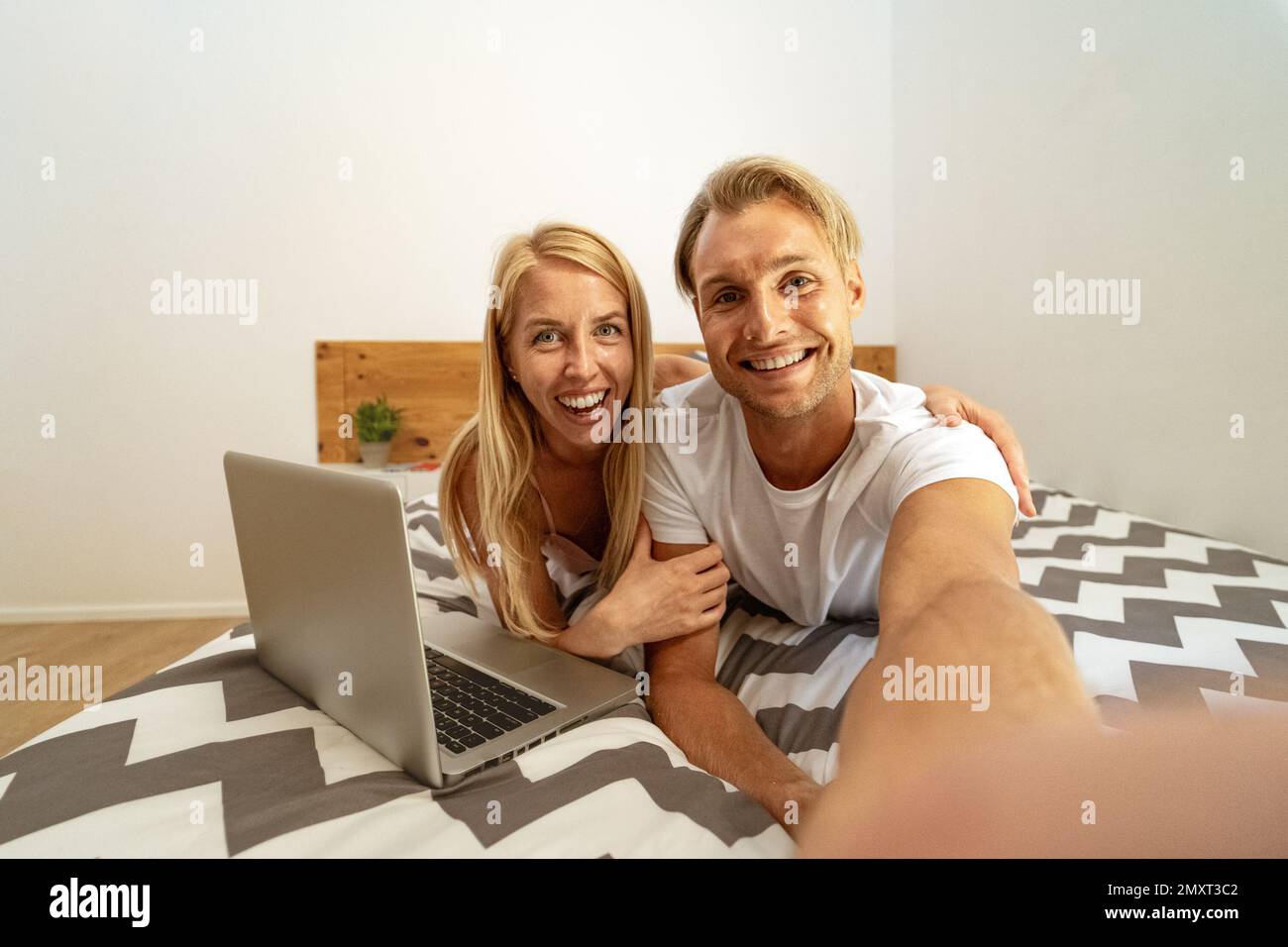Glückliches junges Paar, das Spaß beim Selfie hat, während es ein Notebook im Bett benutzt - Liebes Beziehungs- und Technologiekonzept Stockfoto