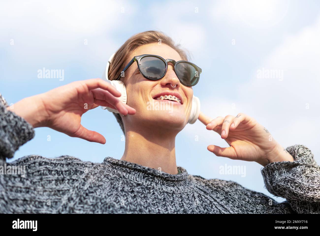 Kleine, kurzhaarige Frau mit Sonnenbrille, die mit großen Kopfhörern Musik hört und lächelt. Stockfoto