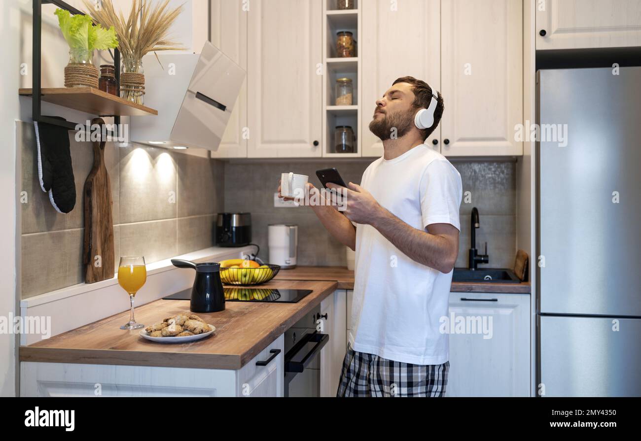 Mann mit Kopfhörern steht in der Küche, trinkt Kaffee und hört Musik und genießt die Morgenzeit. Stockfoto