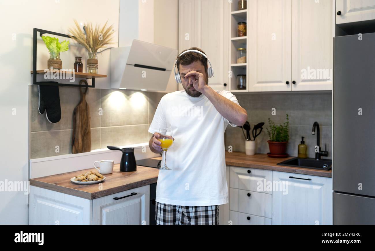 Ein schläfriger junger Mann mit Kopfhörern, der morgens in der Küche steht, mit einem Glas Orangensaft und die Augen mit den Händen reibt. Stockfoto