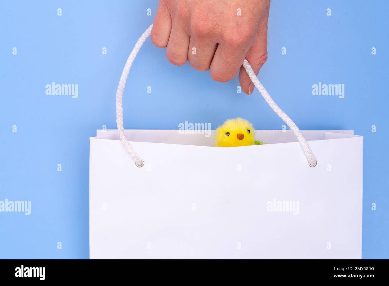 Hand mit einer weißen Papiertüte mit einem gelben, flauschigen Huhn auf blauem Hintergrund. Ostern einkaufen. Konzept Ostergeschenke, Vorbereitung, Urlaub Stockfoto