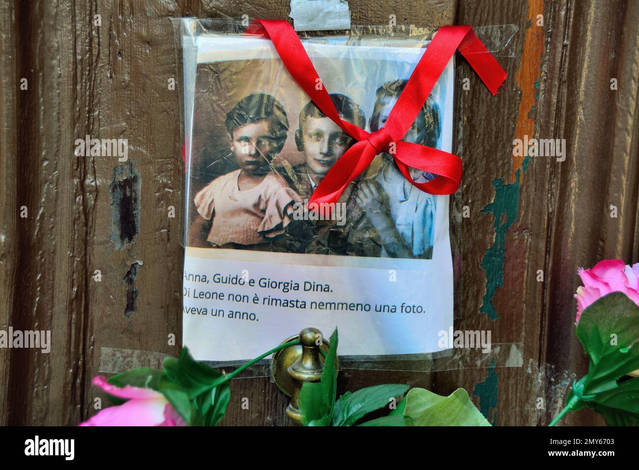 Am Holocaust-Gedenktag 2023 steckte eine anonyme Hand das Bild von drei Kindern und ein paar Blumen an die alte Tür, Nummer 5999 Cannaregio, in Venedig. Die Kinder wurden nach Auschwitz deportiert und 1944 zusammen mit ihrer Großmutter und ihrem einjährigen Bruder getötet. Sie waren 14, 10, 7 Jahre alt. Vor einigen Jahren wurden 7 „Steine“ am Eingang ihres Hauses platziert. Auf dem Foto steht: " Anna, Guido und Giorgia Dina. Leone war ein Jahr alt und wir werden nie wissen, welches Gesicht er hatte. Stockfoto