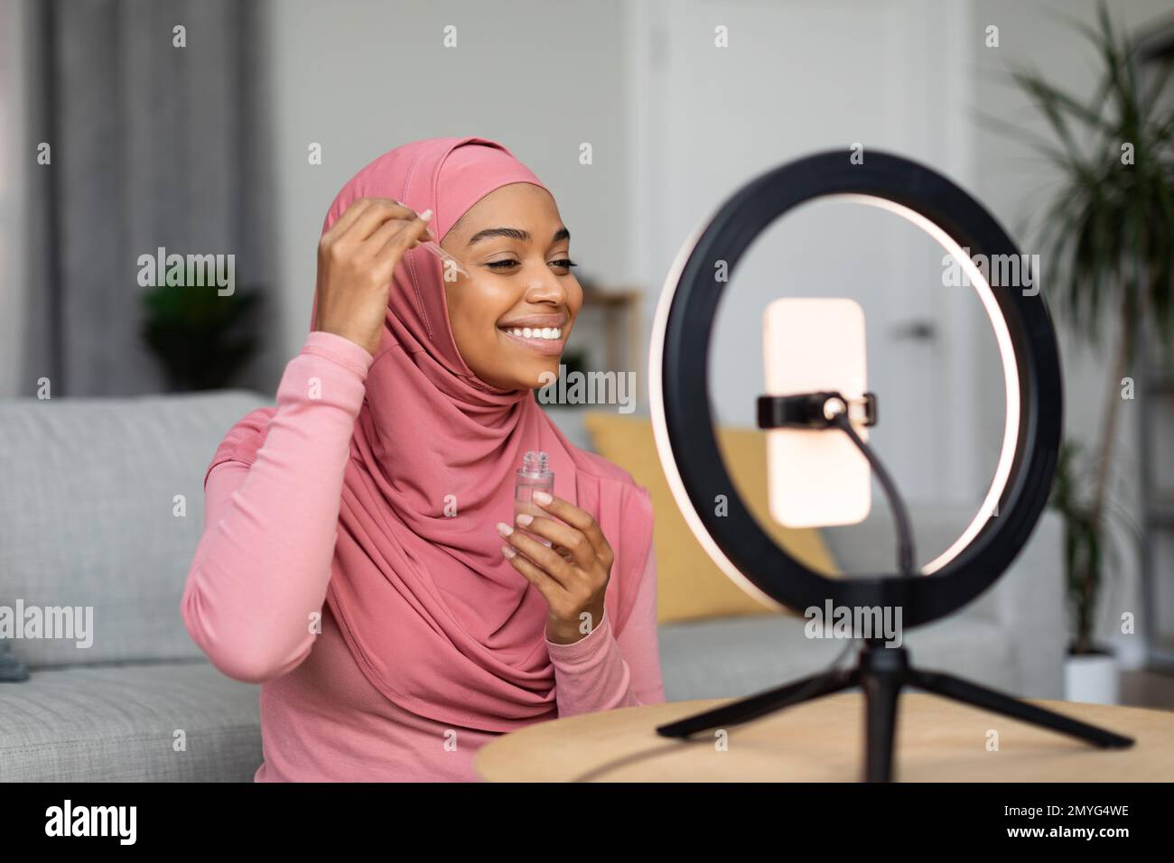 Fröhliche, schwarze, muslimische Schönheitsbloggerin, die ein Video auf einem Smartphone macht, auf einer Ringlampe, die ein neues Gesichtsserum zeigt Stockfoto