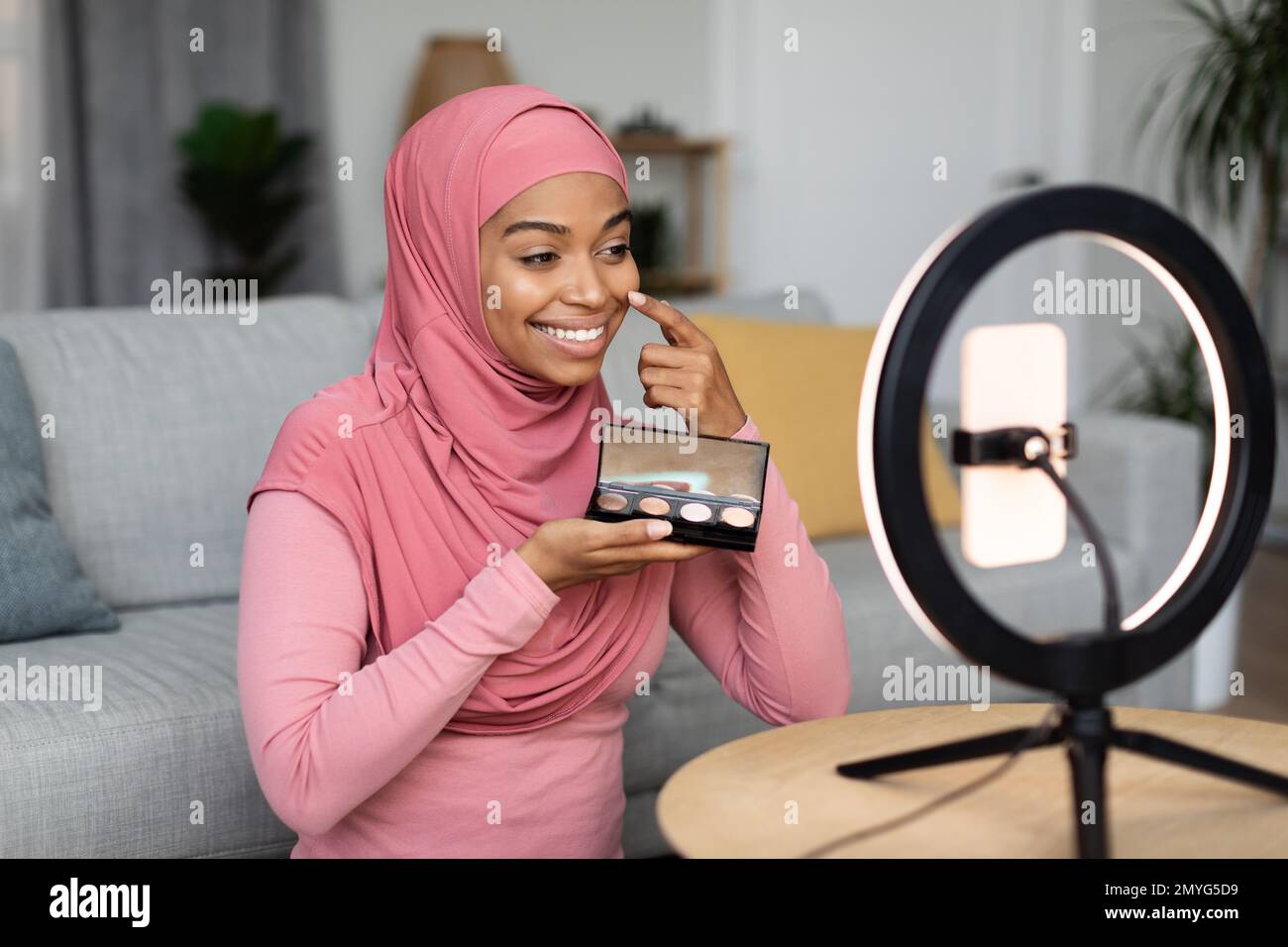 Eine glückliche schwarze moslem-Make-up-Künstlerin sitzt vor einem Handy, das auf einer Ringlampe sitzt, und strömt von zu Hause Stockfoto