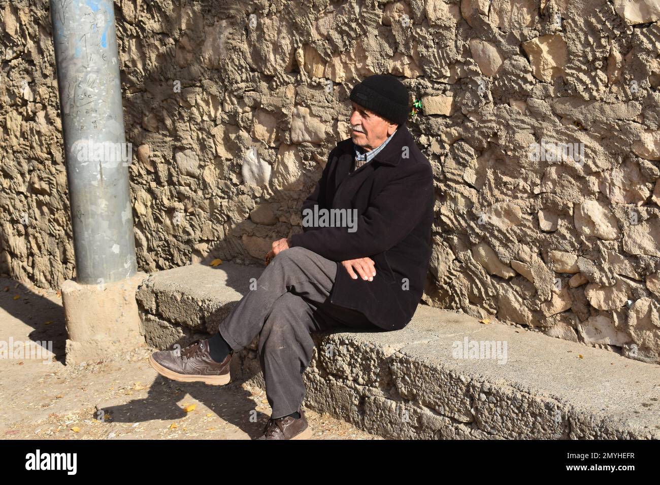 Ein älterer Mann beobachtete die Aktivitäten seiner nordirakischen Stadt von seinem Sitz auf einer Zementbank, die sich gegen eine Steinmauer lehnte. Stockfoto