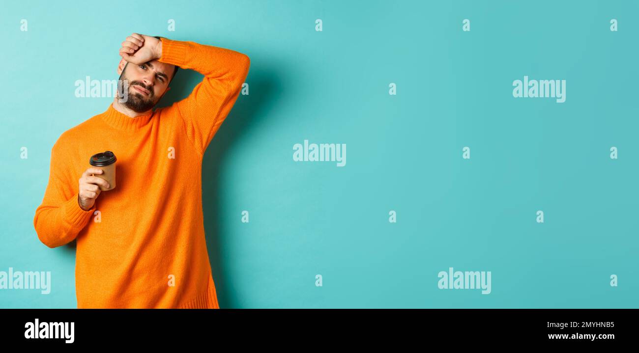 Müde Erwachsene Mann Pause von der Arbeit und Kaffee trinken, wischen Schweiß von der Stirn und sieht erschöpft, in orange Pullover über türkis stehen Stockfoto