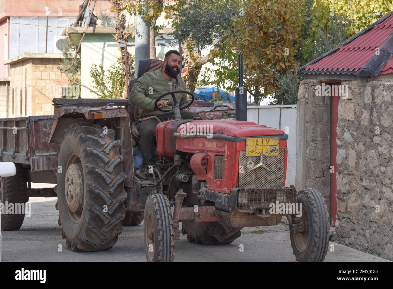 Ein lächelnder Mann, ein Handy in der Hand, führt seinen alten, roten Traktor, der einen Anhänger zieht, durch die Straßen eines Dorfes im Nordirak. Stockfoto