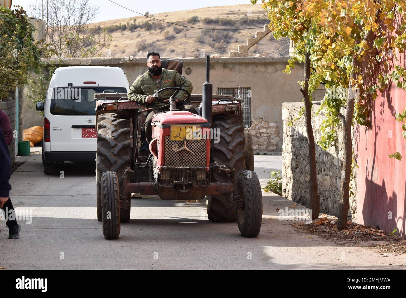 Ein bärtiger Mann führt seinen alten roten Traktor durch die Straßen eines Dorfes im Nordirak. Stockfoto