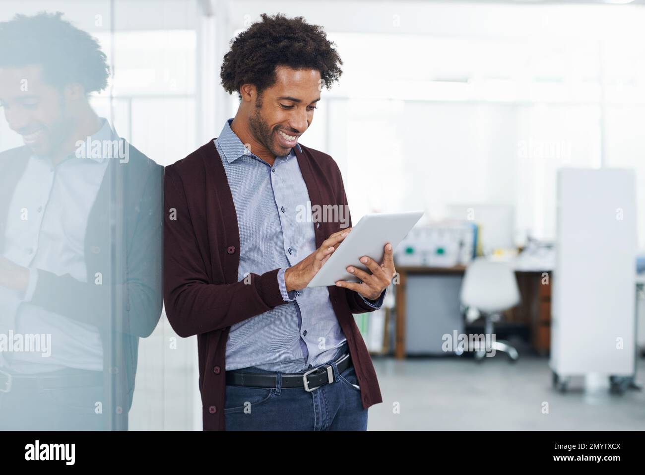 Ich nehme mir Zeit auf der Arbeit. Ein junger Mann lehnt sich an einen Glastrenner, während er ein digitales Tablet benutzt. Stockfoto