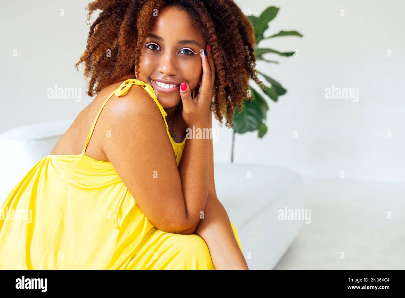 Wunderschöne, kurvige, übergroße junge afto amerikanische Frau in einem gelben Kleid, die zu Hause auf dem Sofa sitzt. Körperpositive, stilvolle, lächelnde Frau hat eine entspannte Zeit. Stockfoto