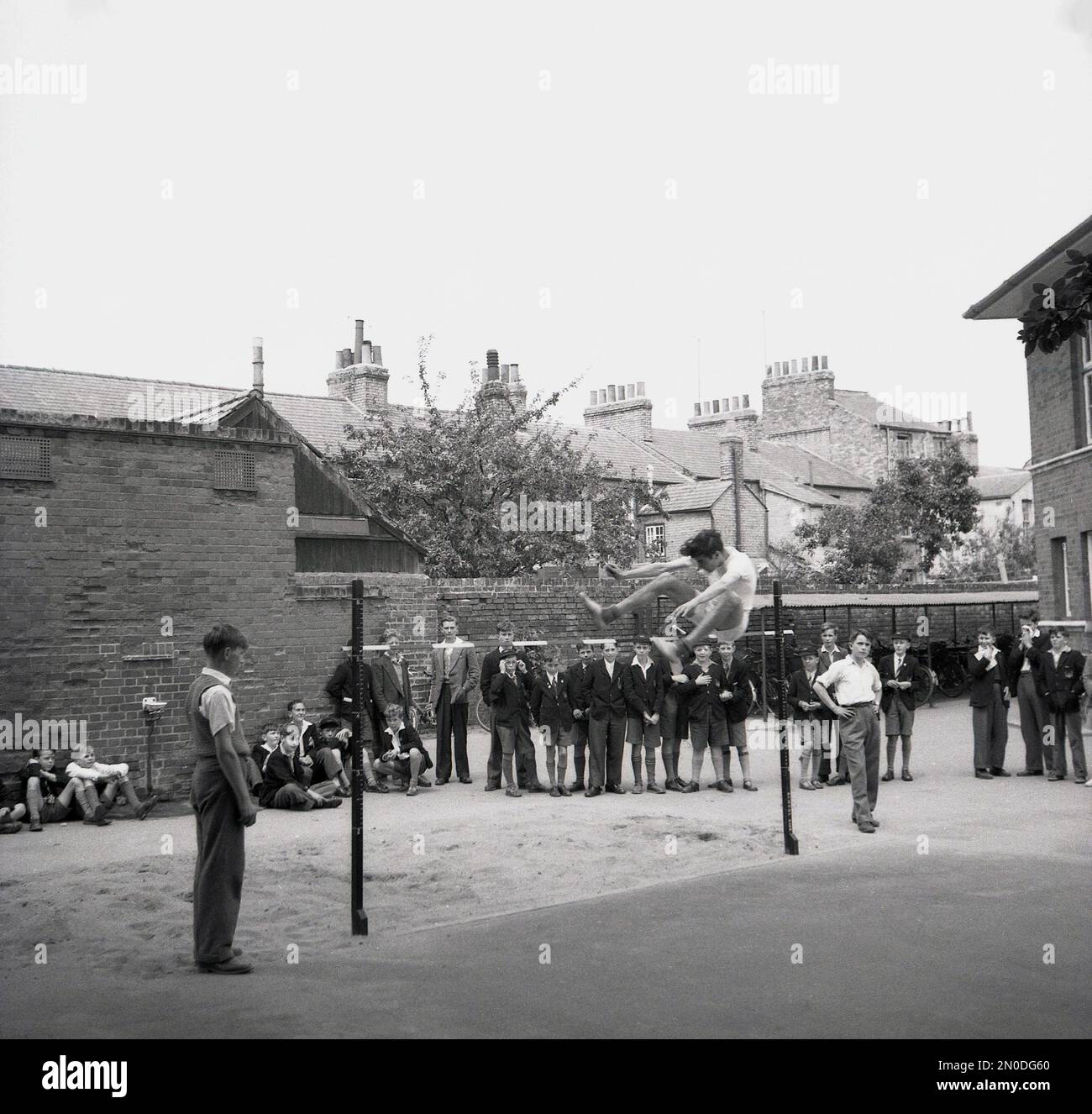 1950er, historisch, draußen auf einem von Mauern umgebenen Schulspielplatz, beobachten eine Gruppe von Schuljungen einen Schuljungen, der in einem High-Jump-Wettbewerb, England, mit den Füßen zuerst über eine Bar springt. Stockfoto