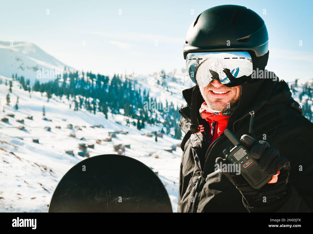 Ein weißer Mann in schwarzer Jacke und Schutzbrille mit Skihelm auf dem Kopf hält ein Walkie-Talkie in der Hand vor dem Hintergrund von weißem Schnee Stockfoto
