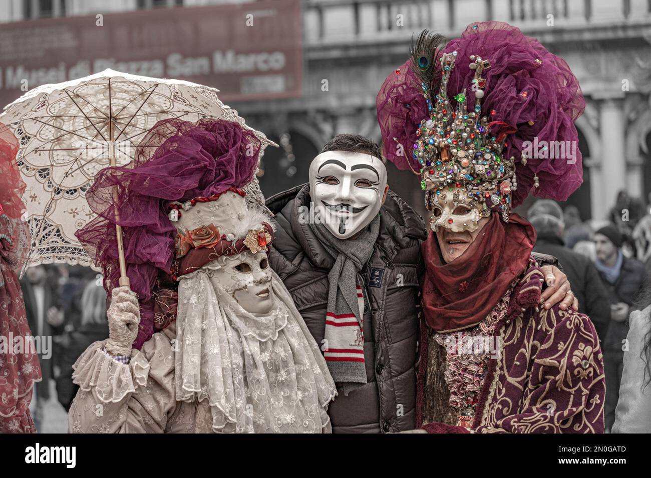 Karneval In Venedig. Ein Mann mit anonymer Maske posiert mit einer kostümierten maskierten Frau mit einem Sonnenschirm und einem kostümierten maskierten Mann mit einem Hut mit Edelsteinen Stockfoto