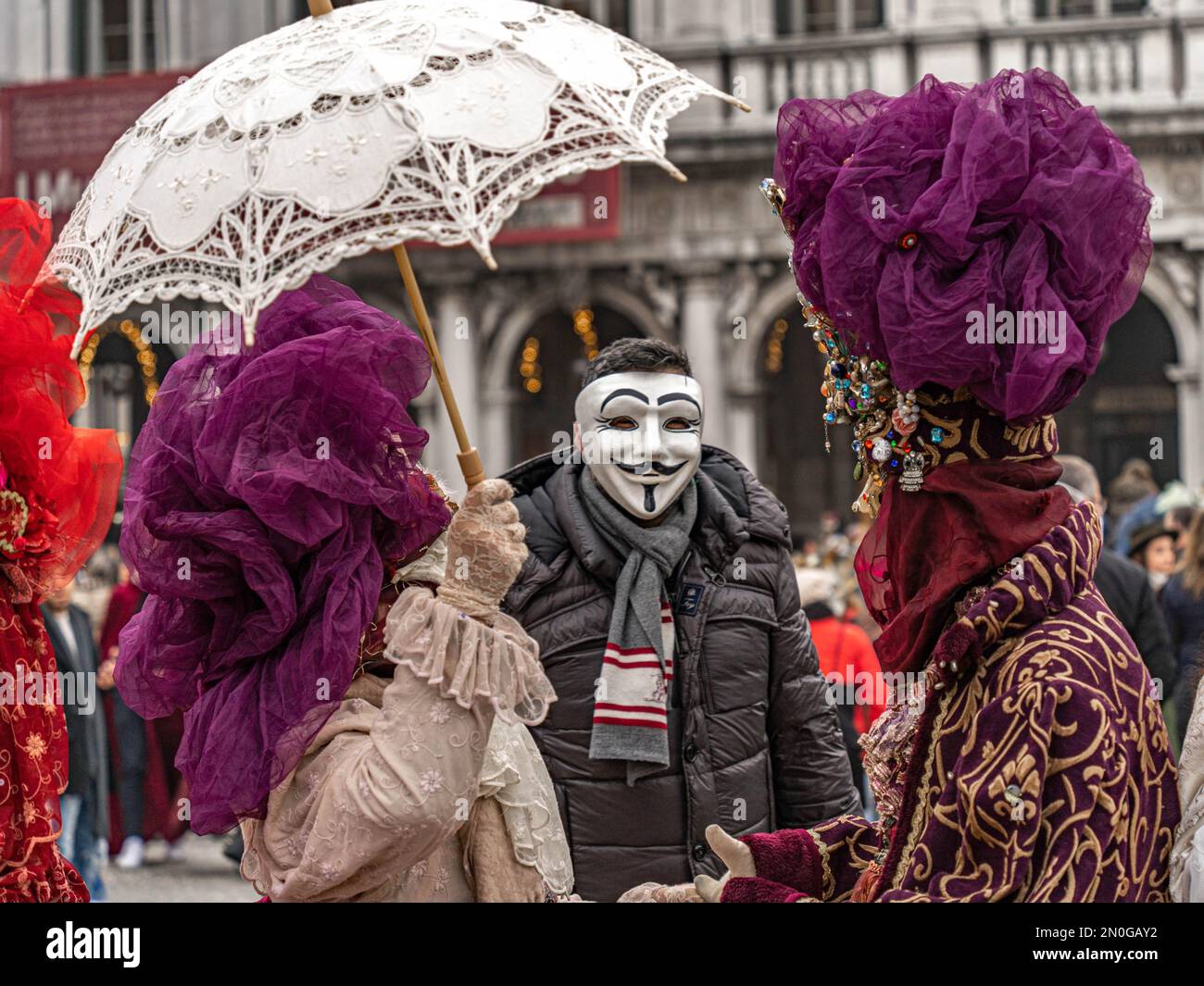 Karneval In Venedig. Ein Mann mit anonymer Maske posiert mit einer kostümierten maskierten Frau mit einem Sonnenschirm und einem kostümierten maskierten Mann mit einem Hut mit Edelsteinen Stockfoto