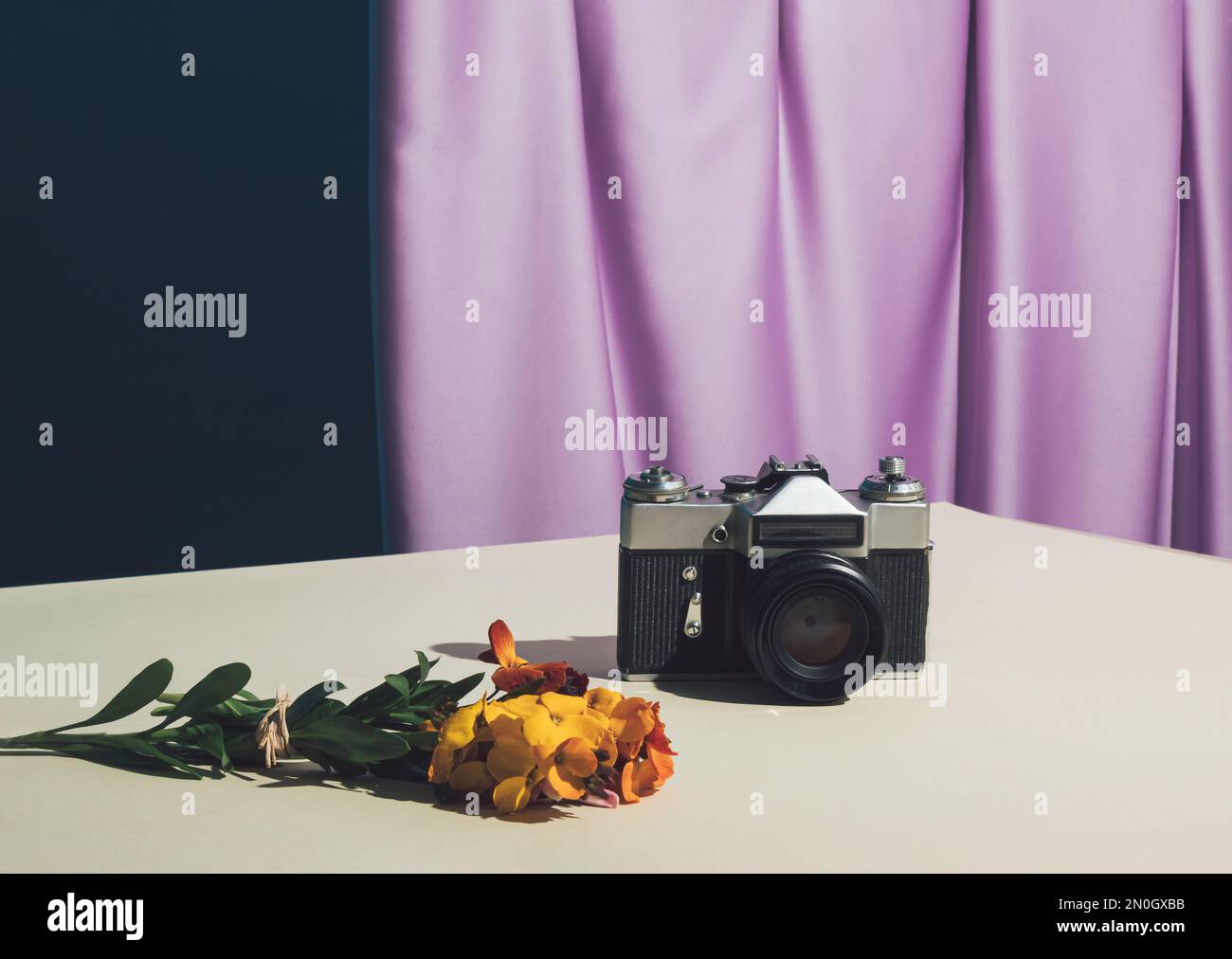 Minimalistisches Konzept mit alter schwarzer Retro-Kamera mit gelben Blumen auf dem Tisch. Hintergrund: Violetter Seidenvorhang. Klassische Komposition. Stockfoto